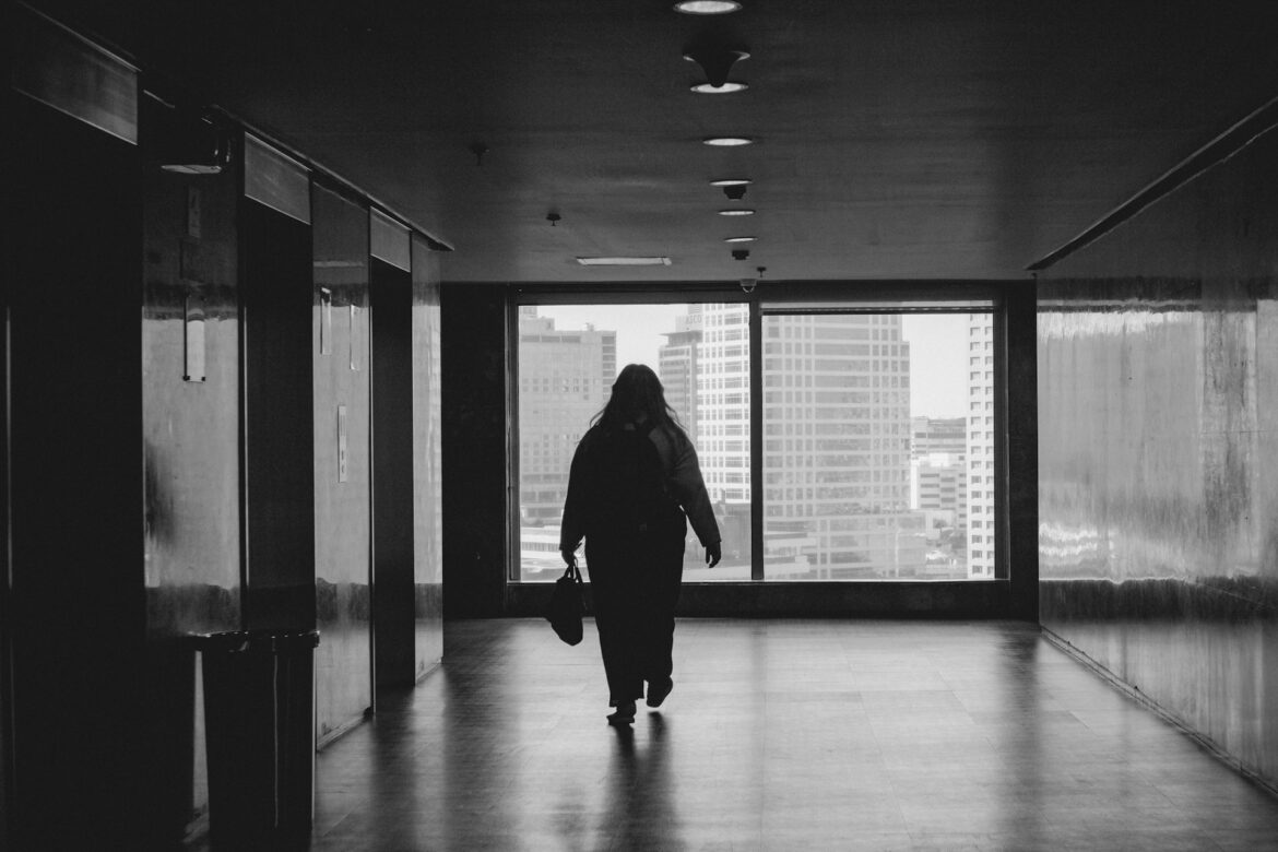 Silhouette of a person walking down a dim hallway toward a large window with city skyscrapers outside.