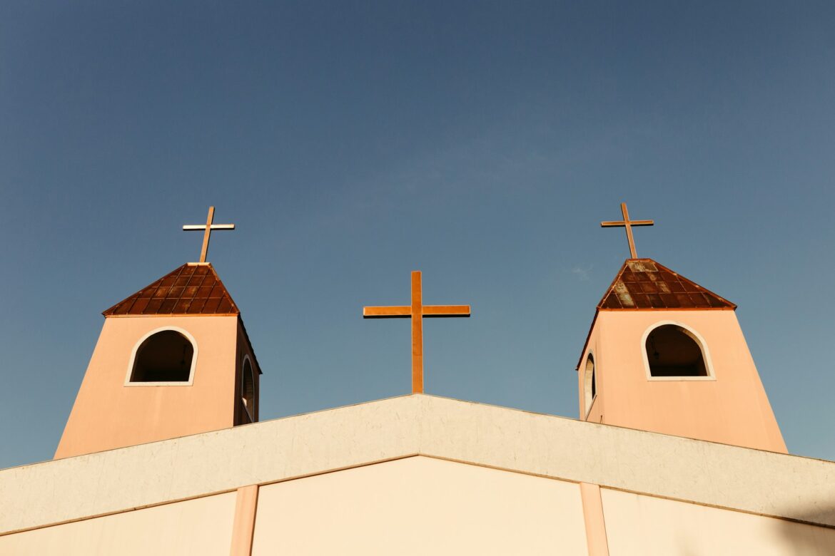 Church facade with two peach-colored towers, each topped by a cross, and a central cross on the roof against a blue sky.