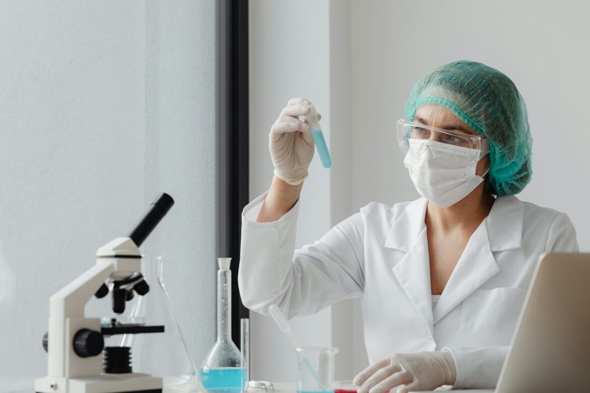 Female scientist in a lab coat, mask, gloves and a hairnet holds a test tube with blue liquid beside a microscope at a lab bench.