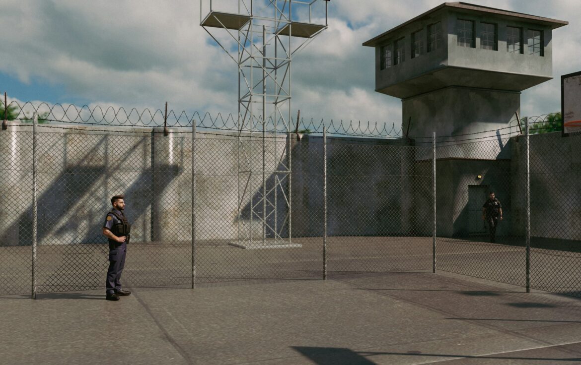 Prison yard with chain-link fencing, barbed wire, and a tall guard tower, with two guards on duty outside the building.