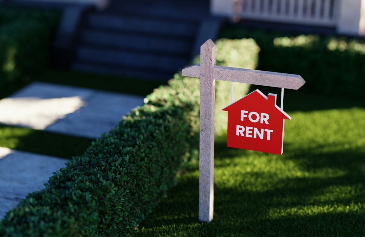 Red 'FOR RENT' house-shaped sign on a wooden post in a well-manicured suburban front yard with a hedge nearby, blue-house steps in the background.